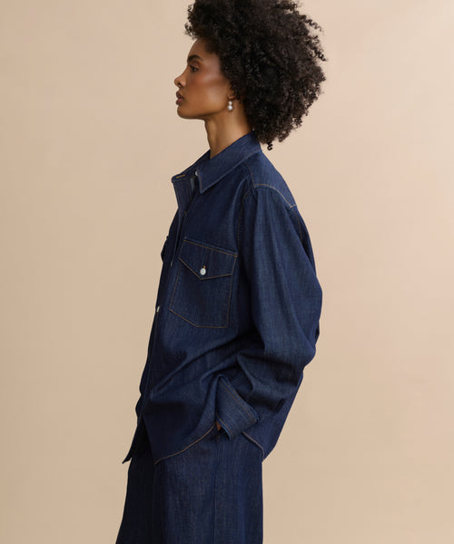 A woman with curly hair stands in profile against a beige background, wearing the Colt Shirt—a Western-inspired dark denim top with buttoned chest pockets—and matching denim pants, hands tucked in her pockets.