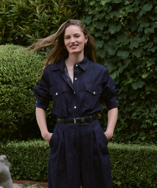 A woman smiles outdoors with her hands in her pockets, wearing the Colt Shirt—a Western-inspired dark denim top paired with matching high-waisted pants. Her long hair is gently windswept as she stands before green bushes.