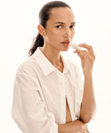 A person with long dark hair in a ponytail, wearing a white button-up shirt, applies Conditioning Lip Balm to their lips against a light background.
