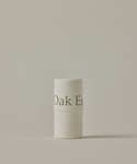 A white cylindrical tube of Conditioning Lip Balm with a minimal design stands upright against a plain beige background. The container features the text Oak Es.