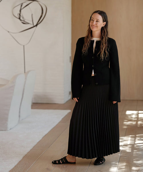 A woman stands indoors on a wooden floor, wearing the Cooper Cardigan over a white top, paired with a long pleated black skirt and black sandals. Sunlight from a window casts patterns while modern art and white furniture appear in the background.