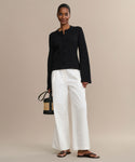 A woman stands against a beige background, wearing the Cooper Cardigan in black with white wide-leg pants and brown sandals. She holds a small woven handbag, smiles gently, and has her hair pulled back.