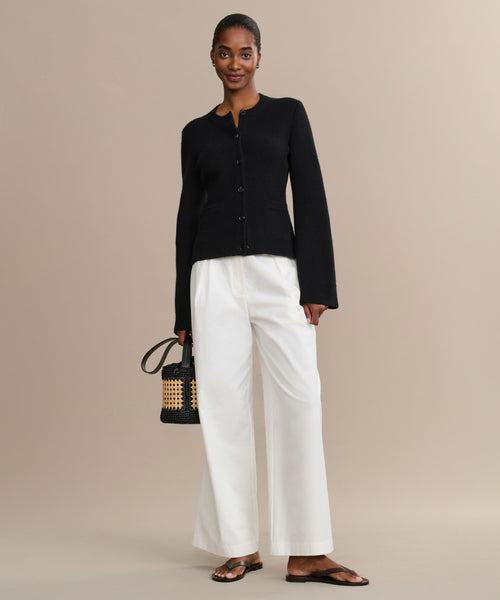 A woman stands against a beige background, wearing the Cooper Cardigan in black with white wide-leg pants and brown sandals. She holds a small woven handbag, smiles gently, and has her hair pulled back.