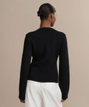 A person with their hair in a neat bun is seen from behind, wearing a fitted black Cooper Cardigan and white pants against a simple beige background.