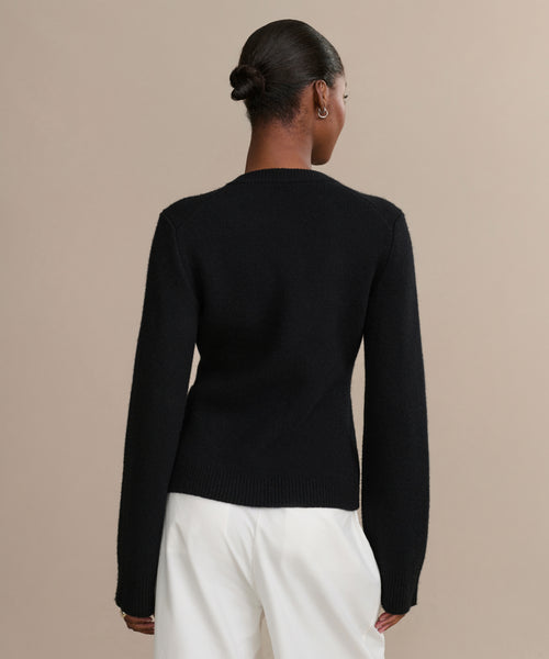 A person with their hair in a neat bun is seen from behind, wearing a fitted black Cooper Cardigan and white pants against a simple beige background.