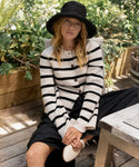 A woman wearing the Cooper Cardigan—a white sweater with ivory and black stripes—paired with a black skirt and white shoes, sits outdoors on a wooden chair surrounded by lush plants and sunlight.