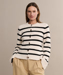 A woman with straight, light brown hair wears the Cooper Cardigan—a cream cardigan with ivory and black stripes, large buttons, and a structured silhouette—paired with beige high-waisted pants against a plain beige background.