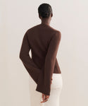 A person stands facing away, wearing the Cooper Cardigan in brown with a structured fit and off-white pants. Their hair is in a neat bun, one hand behind their back, against a plain, light-colored background.