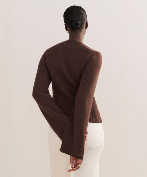 A person stands facing away, wearing the Cooper Cardigan in brown with a structured fit and off-white pants. Their hair is in a neat bun, one hand behind their back, against a plain, light-colored background.