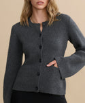 A woman models the Cooper Cardigan, a dark grey button-up with flared sleeves and two front pockets, standing against a beige background. Her left hand is in the pocket, and her face is partially out of frame.