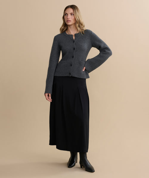 A woman with wavy blonde hair stands against a beige background, wearing the dark grey Cooper Cardigan with a long black skirt and black ankle boots. She gazes slightly to the side, one hand resting on her hip.