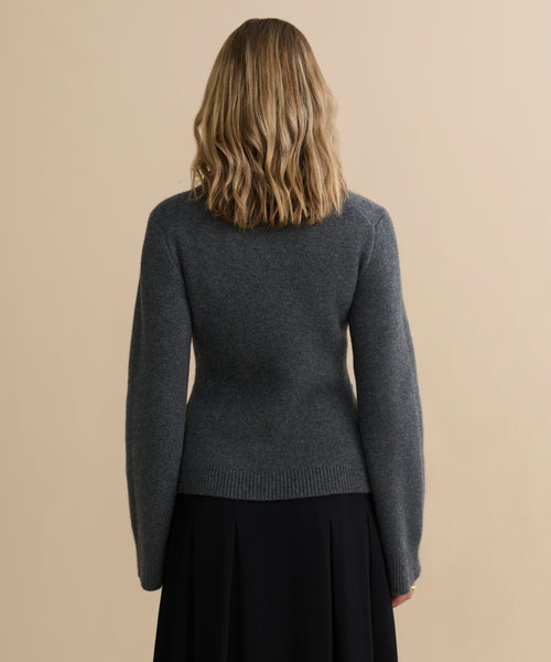 A person with wavy, shoulder-length blonde hair stands facing away from the camera, wearing the fitted dark grey Cooper Cardigan and a black skirt against a plain beige background.