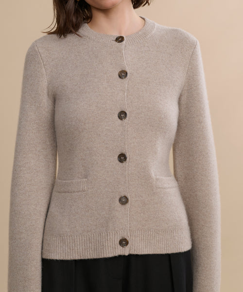 A woman models the Cooper Cardigan—a light beige, long-sleeve, button-up cardigan with a structured fit, front pockets, ribbed hem, and dark buttons—against a plain beige background, with her head partially out of frame.
