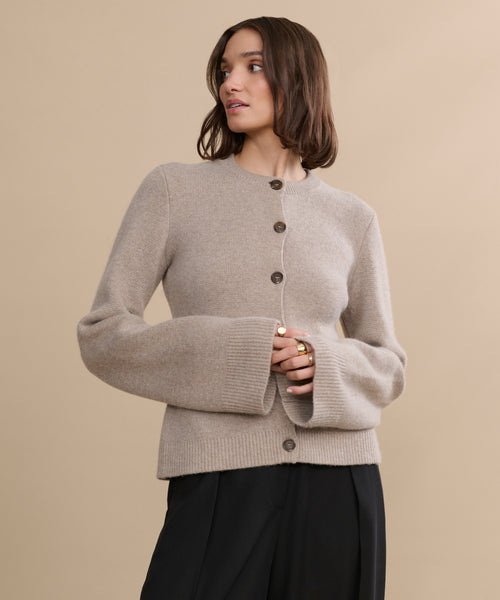 A woman with shoulder-length brown hair models the Cooper Cardigan, featuring wide sleeves and a structured silhouette, paired with black pants, against a plain beige background while looking to her left.