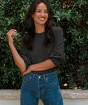 A woman with long dark hair, smiling and looking to the side, stands outdoors in front of green bushes wearing blue jeans and a Boyfriend Long-Sleeve Tee.