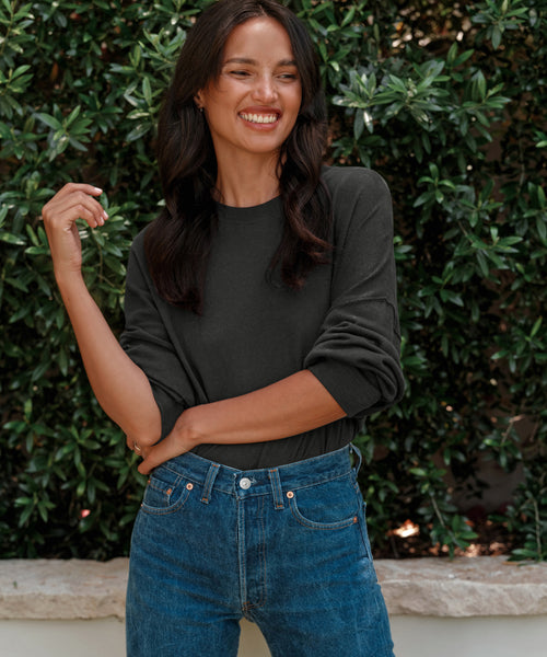 A woman with long dark hair, smiling and looking to the side, stands outdoors in front of green bushes wearing blue jeans and a Boyfriend Long-Sleeve Tee.