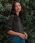A woman with long dark hair models the Boyfriend Long-Sleeve Tee in black and blue jeans, standing outdoors before green leafy bushes and gazing at the camera with a neutral expression.