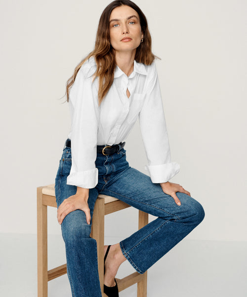 A woman with long brown hair sits on a wooden stool against a plain light background, wearing the Boyfriend Shirt in white and blue jeans, looking directly at the camera with a neutral expression.