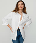 A woman with long brown hair models the oversized Boyfriend Shirt, slightly unbuttoned, paired with blue jeans. She poses with her hands on her hips against a plain light background.