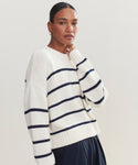 A woman with pulled-back hair wears the Chloe Crewneck, a navy and white striped knit sweater, paired with navy pants. She stands against a plain light background, looking calmly at the camera with her hand near her chin.