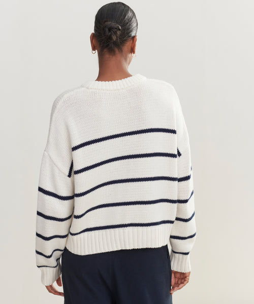 A person with hair in a low bun, wearing the Chloe Crewneck— a white Peruvian cotton sweater with navy stripes— and dark pants, stands facing away from the camera against a neutral background.