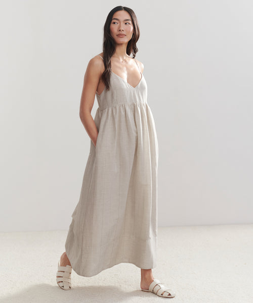 A woman with long hair stands in a bright room, embodying effortless style in the sleeveless, ankle-length Cove Dress with a V-neckline and pockets. She pairs it with white sandals against a minimal background.
