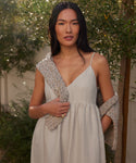A woman with long dark hair stands outdoors near green foliage, wearing a light, sleeveless Cove Dress with a textured, light-colored sweater draped over her shoulders.