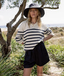 A woman stands outdoors amid greenery and a glimpse of the sea, wearing the Cotton Kate V-Neck striped sweater with navy accents, a wide-brimmed hat, and dark shorts.