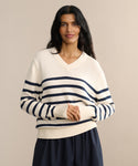 Woman with long dark hair wears the Cotton Kate V-Neck, a cream-striped cotton sweater, paired with a navy skirt against a beige backdrop—smiling with hands clasped for an ideal spring knit look.