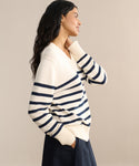 A woman with long dark hair, wearing the Cotton Kate V-Neck striped sweater and navy pants, stands in profile with her hand touching her chin, smiling slightly against a plain beige background.
