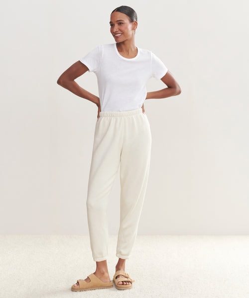 A woman stands smiling with hands on hips, wearing an ultra-soft white t-shirt, Saturday Sweatpant joggers in cream, and beige slide sandals against a light neutral background.