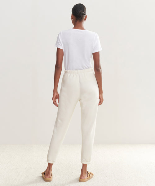 A woman stands with her back to the camera wearing a plain white T-shirt, Saturday Sweatpant joggers in ultra-soft French terry, and tan moccasin slippers against a light, neutral background.