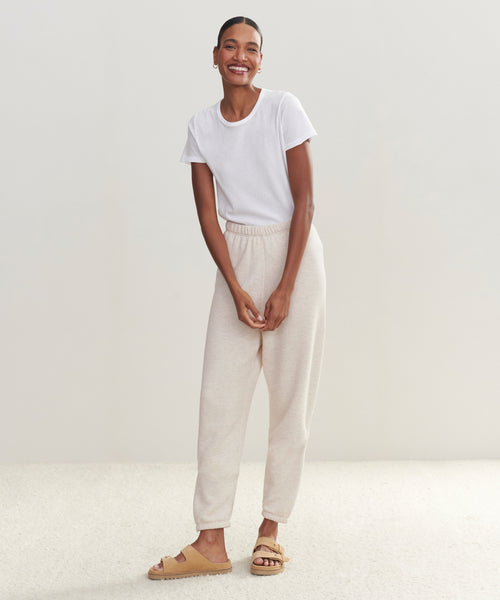 A smiling woman stands against a plain light background, wearing a white t-shirt and the ultra-soft Saturday Sweatpant joggers. She pairs them with tan slide sandals, has short hair, and rests her hands in her pockets.