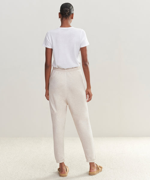 A person stands indoors facing away, wearing a white t-shirt, ultra-soft light beige Saturday Sweatpant made from French terry, and tan slippers against a plain neutral background.