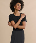 A woman with curly hair wears the Cotton Slim Tee in black and a black pleated skirt, standing against a beige background, smiling and looking to the side with her arms gently crossed.
