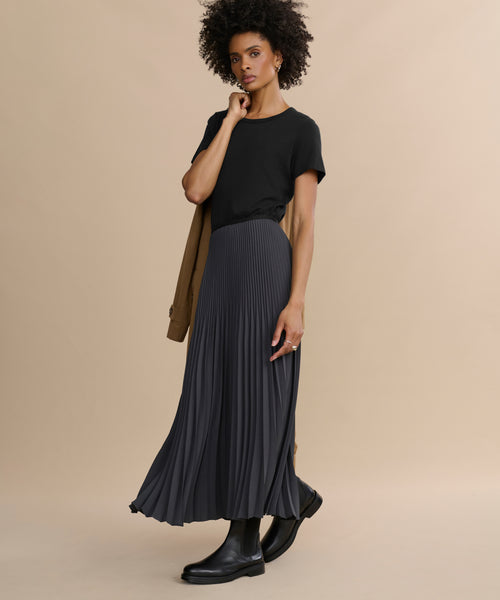 Wearing a Cotton Slim Tee with a fitted silhouette, a long pleated black skirt, and black boots, a woman stands against a beige background holding a camel coat over her shoulder.