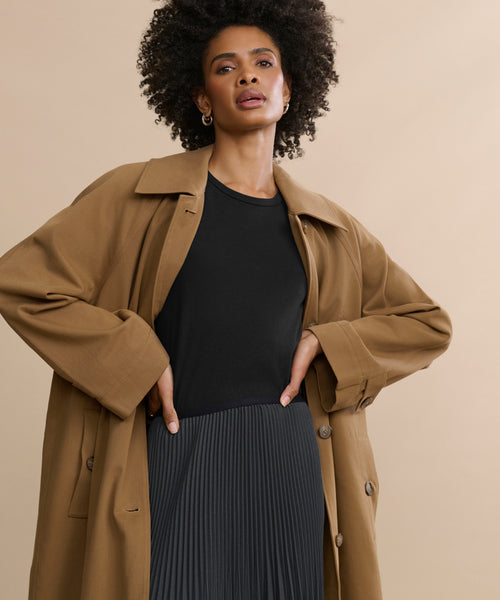 Wearing a tan trench coat over a black Cotton Slim Tee and pleated skirt, a person with curly hair stands against a beige background, hands on hips and looking confidently at the camera.