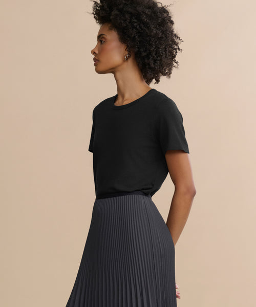 A woman with curly hair stands in profile against a beige background, wearing the Cotton Slim Tee—a black, crewneck, fitted top—paired with a high-waisted, pleated dark skirt.