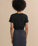 A person with curly hair, seen from behind, wears the Cotton Slim Tee in black, tucked into a high-waisted dark gray pleated skirt, creating a fitted silhouette against a plain beige background.
