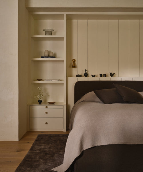 A cozy, minimalist bedroom with a Cove Bed featuring an upholstered headboard, beige built-in shelves for vases and books, a compact nightstand, a dark brown rug, and neatly made Libeco linen bedding in beige and brown.