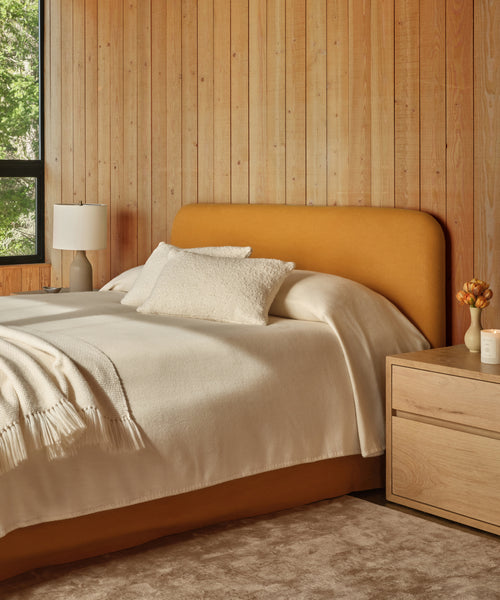 A cozy bedroom with wood-paneled walls features the Cove Bed, which has an upholstered mustard linen headboard, cream bedding, a throw blanket, bedside table with a lamp, flowers in a vase, and a candle. Sunlight streams through the window.