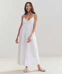 A woman smiles in the Cove Dress—a relaxed fit, sleeveless white dress with thin straps—paired with white sandals. She has long, dark hair and stands in a minimalist room with light-colored floors and walls.