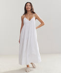 A woman poses against a plain light background wearing the Cove Dress, a long white cotton dress with thin straps and white sandals. Her hair is down, and one hand rests on her hip, showcasing a relaxed fit style.