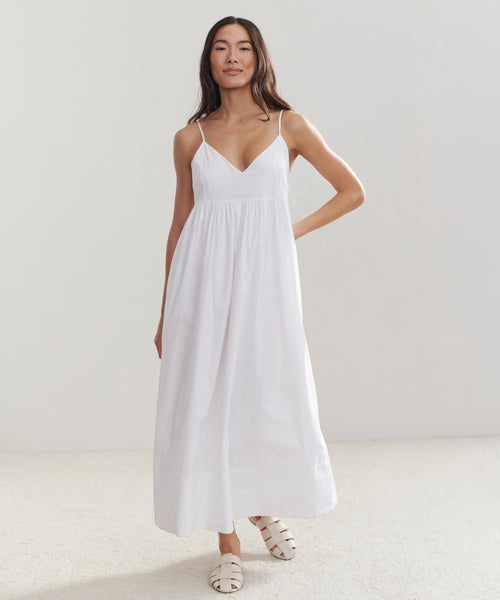 A woman poses against a plain light background wearing the Cove Dress, a long white cotton dress with thin straps and white sandals. Her hair is down, and one hand rests on her hip, showcasing a relaxed fit style.
