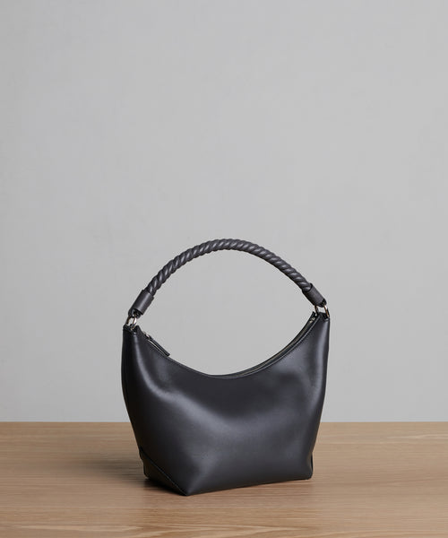 The Crescent Shoulder Bag, crafted in Italy from soft leather with a twisted handle, is displayed on a light wooden surface against a plain gray background.