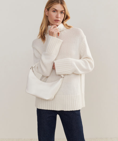 A woman in a cream turtleneck and dark jeans poses with the Crescent Shoulder Bag, crafted in Italy from supple lambskin, stylishly slung over her shoulder against a plain beige backdrop.