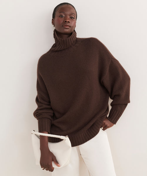 A woman in a loose brown turtleneck and white pants stands against a neutral background, confidently holding the Crescent Shoulder Bag. She has one hand in her pocket and looks directly at the camera.