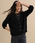 A smiling woman with long dark hair wears the Cropped Cashmere Cocoon Cardigan in black over a matching top and dark jeans, standing against a beige background with one hand on her hip and the other raised above her head.