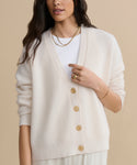 A woman wearing the Cropped Cashmere Cocoon Cardigan in ivory over a white top, accessorized with gold necklaces, a ring, and a bracelet, stands against a beige background.