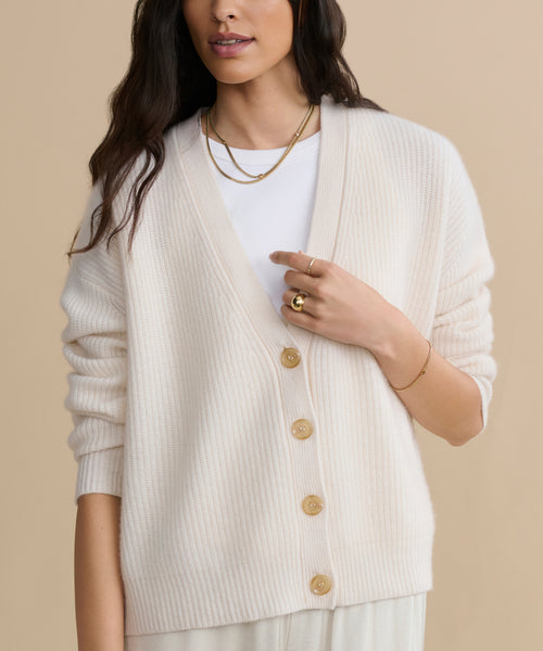 A woman wearing the Cropped Cashmere Cocoon Cardigan in ivory over a white top, accessorized with gold necklaces, a ring, and a bracelet, stands against a beige background.